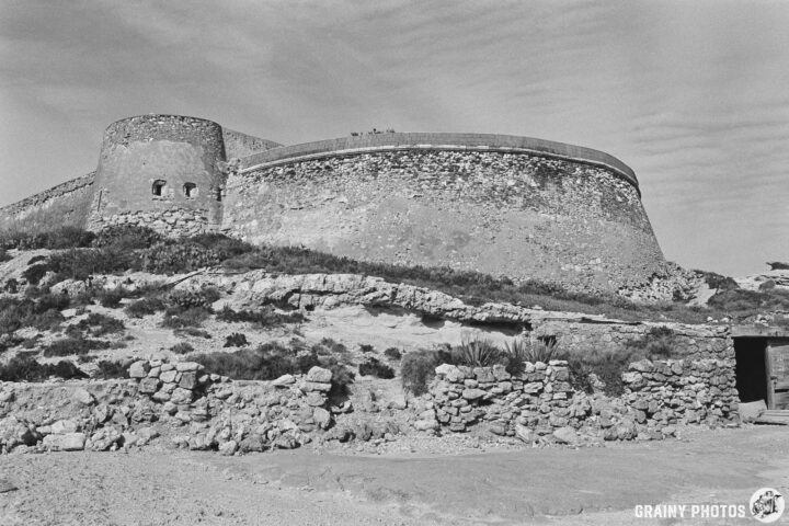 A black and white image of Castillo de San Ramón, featuring a circular turret and surrounding rocky terrain. The fortress stands against a softly textured sky, showcasing its sturdy walls and the natural landscape.