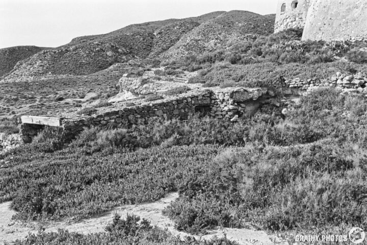 A black and white image capturing a rugged landscape with a stone structure partially hidden by vegetation, with the walls of Castillo de San Ramón surrounded by rolling hills. The scene evokes a sense of history and nature, with remnants of human construction amidst the natural terrain.