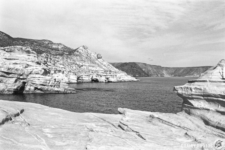 A black and white landscape featuring rocky cliffs and calm water, with mountains in the background under a cloudy sky.