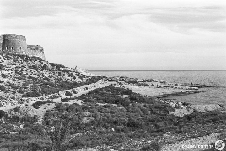 A coastal landscape featuring Castillo de San Ramón and rugged terrain. The scene captures the serene ocean, with a winding path leading to the water’s edge, framed by vegetation under a cloudy sky.