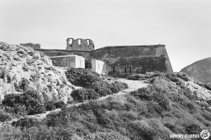 A black and white image of the entrance to Castillo de San Ramón situated on a hill, surrounded by greenery and rocky terrain. The structure features ruins with visible openings and a modern addition, showcasing a contrast between old and new architecture.