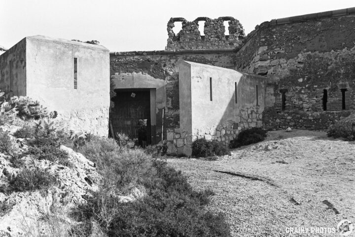 The entrance to Castillo de San Ramón with a wooden door, surrounded by rocky terrain and sparse vegetation. The remnants of an old fortification rise above, featuring broken walls against a stark sky. The image is captured in black and white, emphasizing its historical significance.