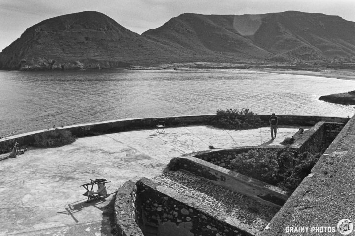 A serene black and white landscape featuring a coastal view from Castillo de San Ramón with rugged mountains in the background, a person standing on a stone terrace, and scattered outdoor furniture, evoking a sense of solitude and natural beauty.