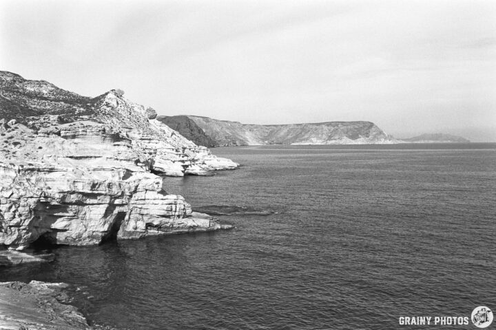 Black and white image of a rocky coastline with cliffs and calm ocean waters. The distant hills outline the horizon under a soft, cloudy sky, creating a serene natural landscape.