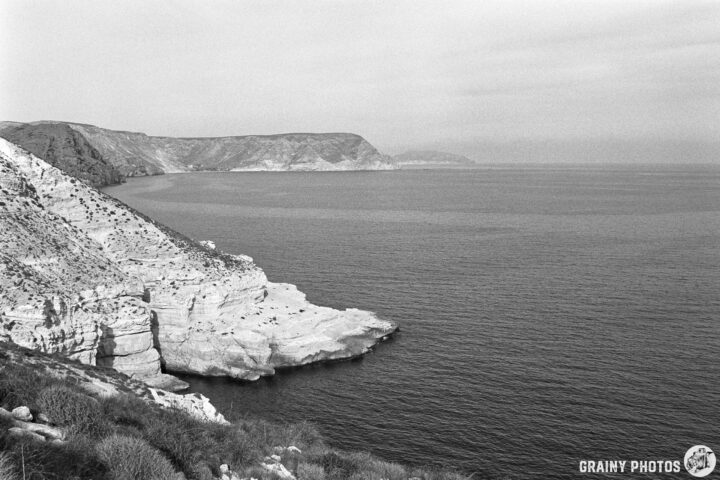 A black and white coastal landscape featuring rugged cliffs that meet a calm sea. The horizon blends softly with the sky, creating a serene atmosphere. The rocky shoreline is dotted with shrubs, adding texture to the natural scenery.