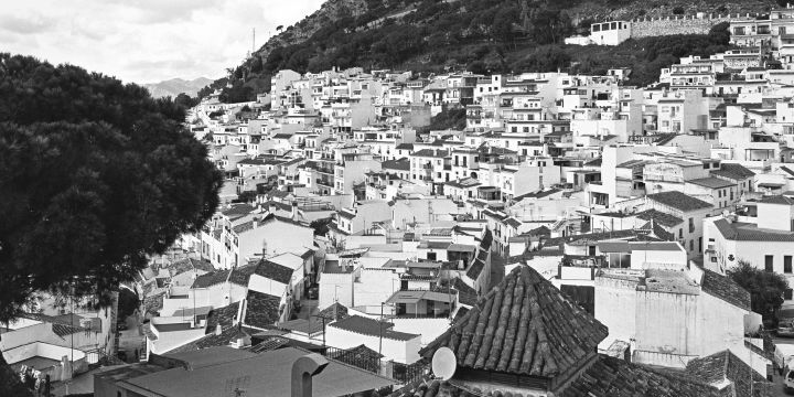 A black and white photograph of Mijas Pueblo with densely packed white buildings, nestled against a mountainous backdrop. The scene captures the charm of traditional architecture and lush greenery in the foreground.