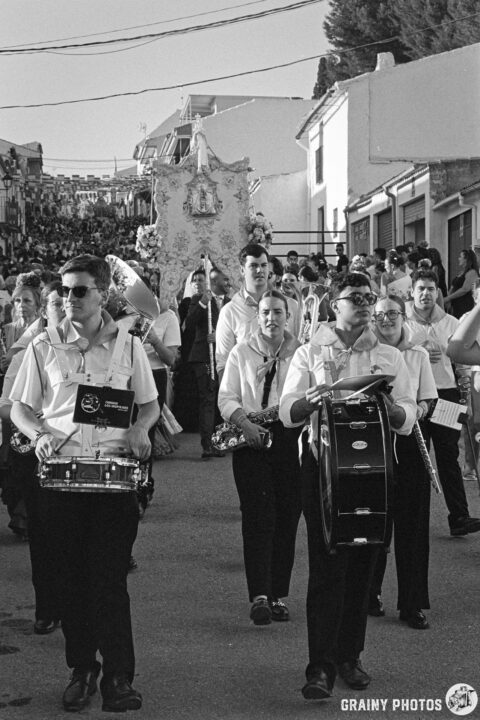 A lively procession featuring musicians in white shirts and black pants, playing drums and marching. A crowd gathers along the street, with decorations and festive energy visible all around.