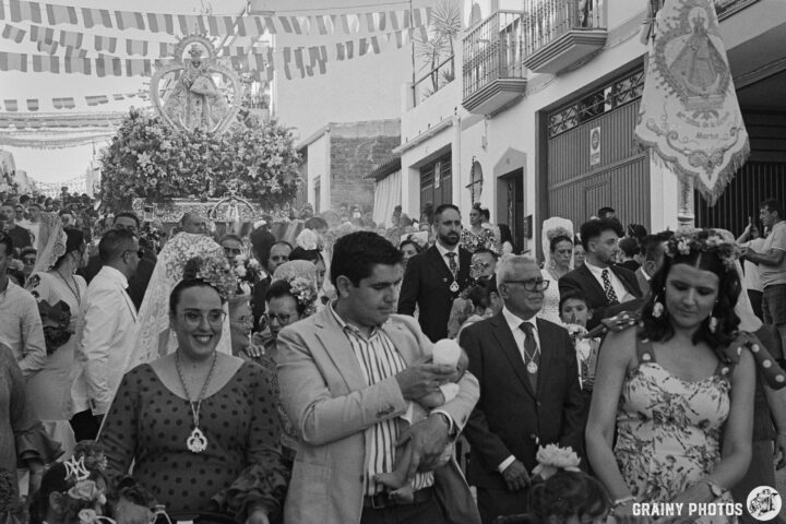 A lively processional scene during a local Romería Virgen de la Victoria de Martos  festival, featuring a crowd dressed in festive attire, flowers in hand, celebrating under colorful decorations. The Main Throne with a Figure of the Virgen de la Victoria de Martos is visible in the distance