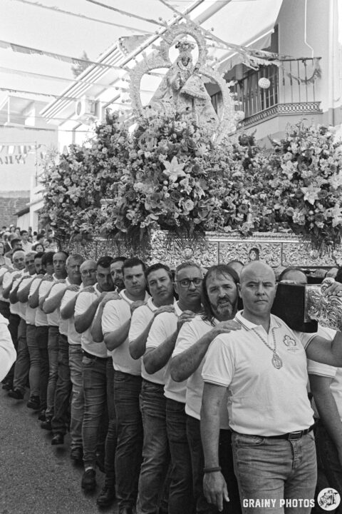 A procession featuring a group of men in matching white shirts carrying a floral-adorned throne with a statue of the Virgen de la Victoria de Martos. They maintain a serious demeanor while marching down a street decorated for a celebration.