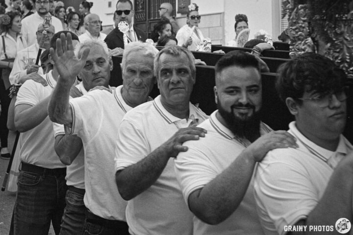 A closeup photo of a group of the throne bearers wearing white polo shirts, with some waving and smiling. The scene is lively, featuring a diverse crowd in the background, contributing to a festive atmosphere, captured in black and white.