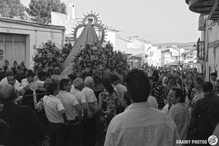 A lively street celebration featuring a procession with a floral-adorned statue of the Virgen de la Victoria de Martos, surrounded by a crowd of people in traditional clothing, amidst white buildings and trees in the background, capturing the essence of a cultural festival.