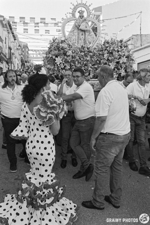 A lively street scene during a festival, featuring a woman in a polka-dot flamenco dress talking to some of the throne bearers standing next to the flower adorned float with a statue of the Virgen de la Victoria de Martos. The atmosphere is vibrant and festive, capturing a cultural celebration.