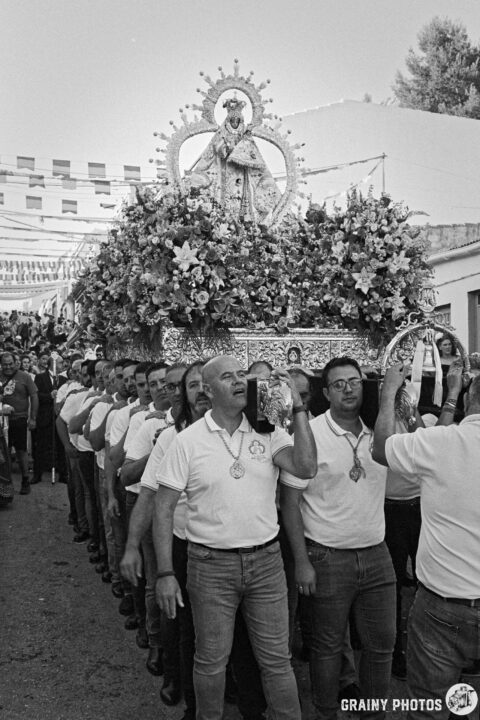 A procession featuring a statue of the Virgen de la Victoria de Martos adorned with flowers, carried by a group of men in white shirts. The atmosphere is festive, with decorations and a crowd in the background, celebrating a local festival or religious event.
