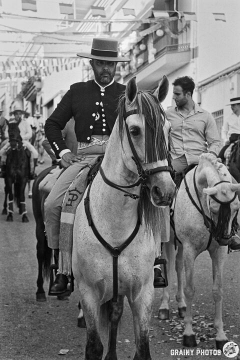 A man dressed in traditional Spanish attire rides a white horse during a festive street event. The scene captures vibrant decorations and a lively atmosphere, with other horse riders in the background, emphasizing the cultural celebration.