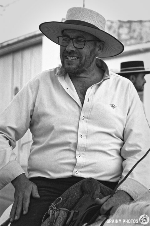 A man wearing a wide-brimmed hat and glasses smiles while seated on a horse. He is dressed in a light-colored shirt and dark trousers, with another man in the background wearing a similar hat. The image is in black and white, capturing a rural or traditional atmosphere.