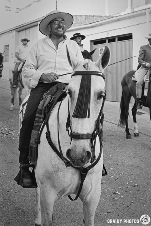 A smiling man in a white shirt and hat rides a white horse, leading a group of horseback riders down a street. The scene is in black and white, capturing a lively atmosphere during a festive event.