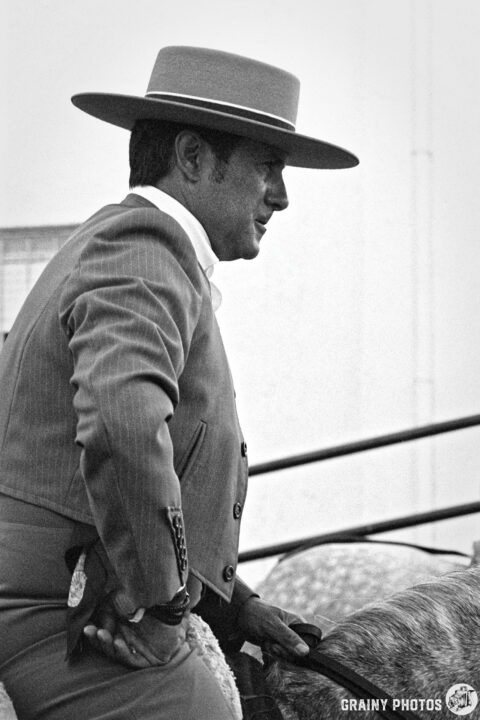 A man dressed in traditional equestrian attire, wearing a wide-brimmed hat, sits atop a horse. The image is in black and white, capturing a moment of concentration and elegance at the Romería Virgen de la Victoria de Martos.