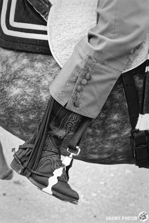 Close-up of a rider's leg in a grayish saddle with decorative buttons, wearing ornate cowboy boots with fringes. The background features a horse, emphasizing the equestrian theme in a monochromatic setting.