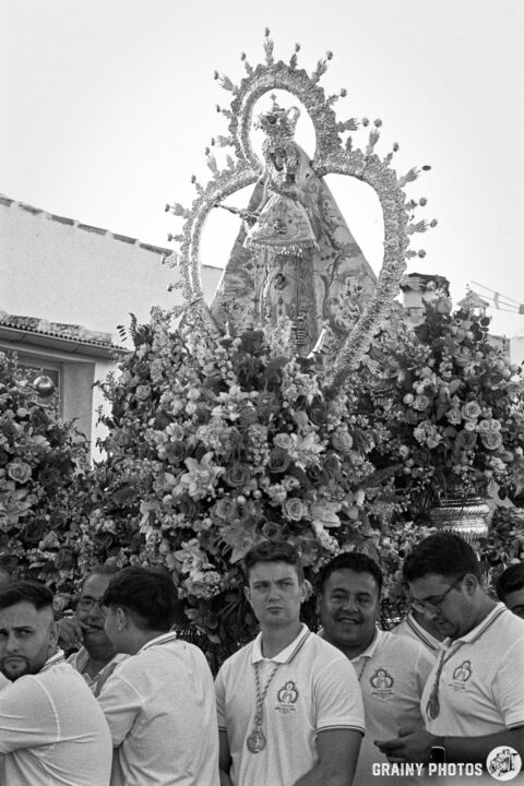 A group of men in matching white shirts stands amid vibrant flower arrangements, carrying a decorative religious statue. The atmosphere suggests a traditional procession or celebration, highlighted by the intricate details of the statue and floral displays.