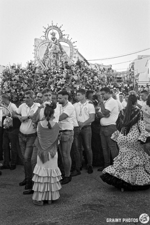 A procession with a floral-adorned statue at its center, surrounded by people in traditional attire. Some wear white shirts while others don colorful dresses, celebrating a cultural event filled with community spirit and tradition.
