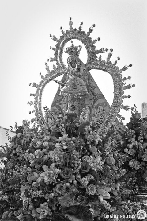 A statue of the Virgen de la Victoria de Martos, adorned with a crown and surrounded by intricate floral arrangements, featuring a mix of roses and other flowers, set against a clear bright sky.