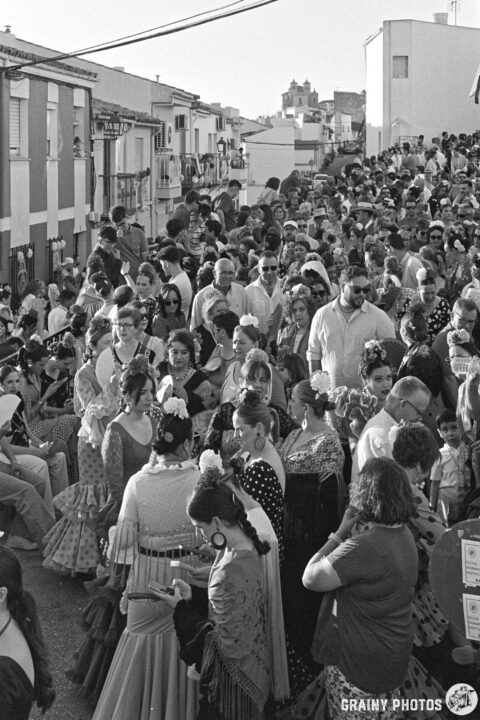 A vibrant street Romeria scene in black and white, filled with a large crowd of people dressed in traditional attire, celebrating together. The atmosphere is lively with a mix of music and cultural expressions.