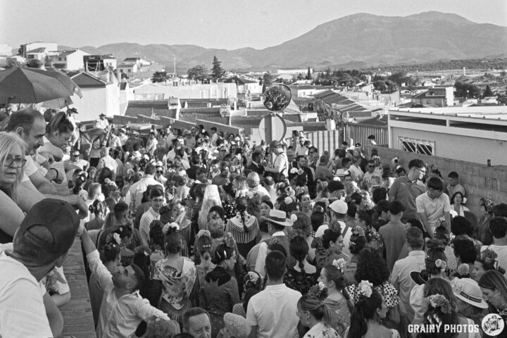 A crowded gathering at the Martos Romeria, featuring people in traditional attire and floral accessories. The backdrop includes rooftops and distant mountains, indicating a festive atmosphere. The image is in black and white, highlighting the lively scene.