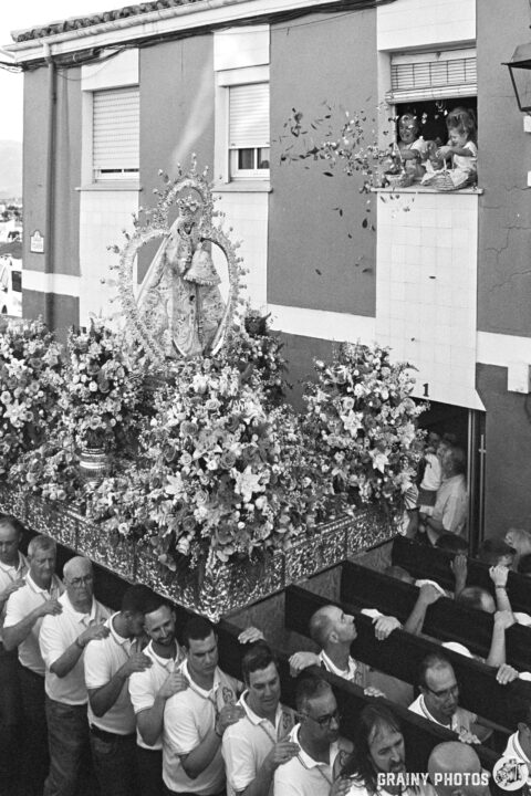 A procession featuring a decorated throne with a statue of the Virgen de la Victoria de Martos adorned in flowers, accompanied by a group of men carrying it. Onlookers watch from a window above throwing flower petals. Festive decorations and a sense of community celebration in a vibrant street setting.