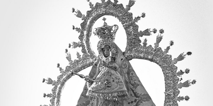 A statue of the Virgen de la Victoria de Martos, adorned with a crown and surrounded by intricate floral arrangements, featuring a mix of roses and other flowers, set against a clear bright sky.